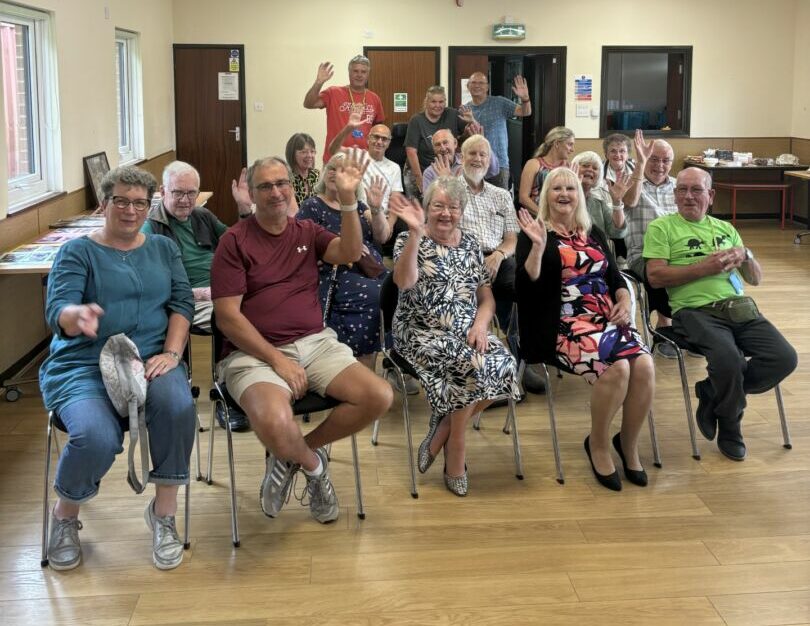 The Lancashire and Cumbria group sitting in chairs, smiling and waving in a welcoming fashion towards the camera.